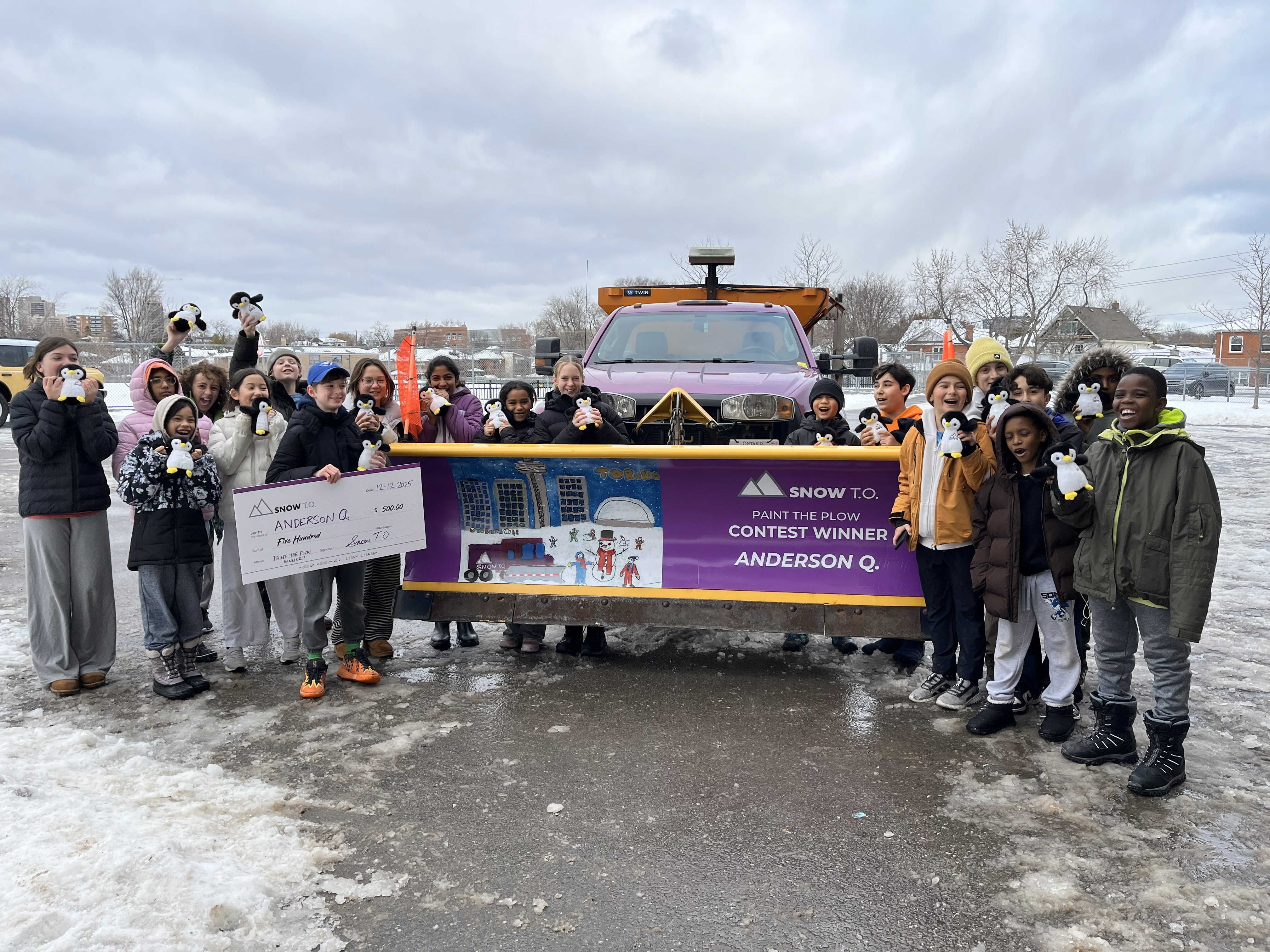 Group of students stands in a parking lot on either sides of a snow plough, each student holding a stuffed penguin smiling