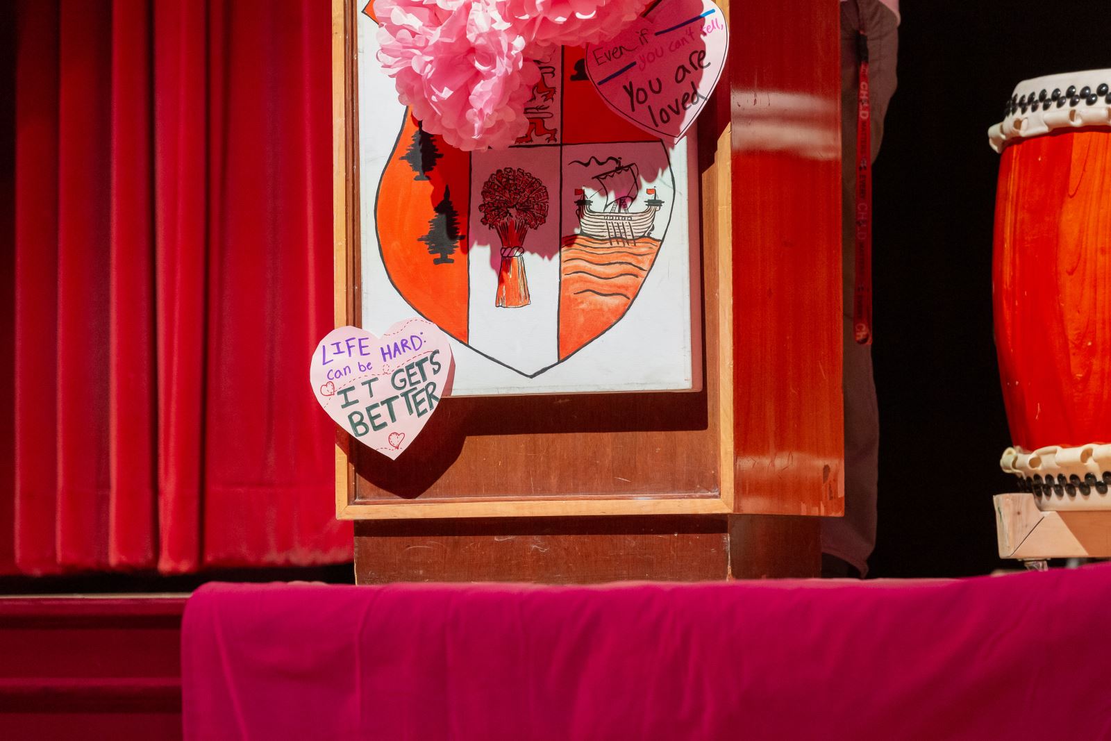 A picture of a base of a podium at the International Day of Pink Event at Harbor CI, showing off a cut out heart that says Life Can Be Hard, It Gets Better.