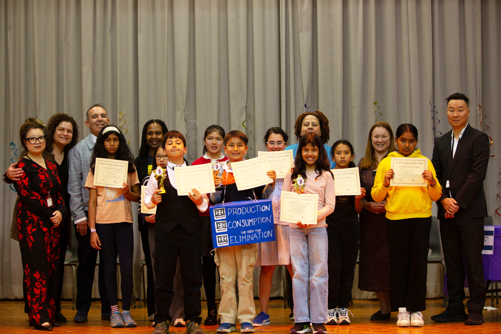 A group of students and adults stand on a stage in front of a grey curtain at an awards event. Several students in the front row hold Toronto District School Board certificates and star-shaped trophies, while adults stand behind them. 