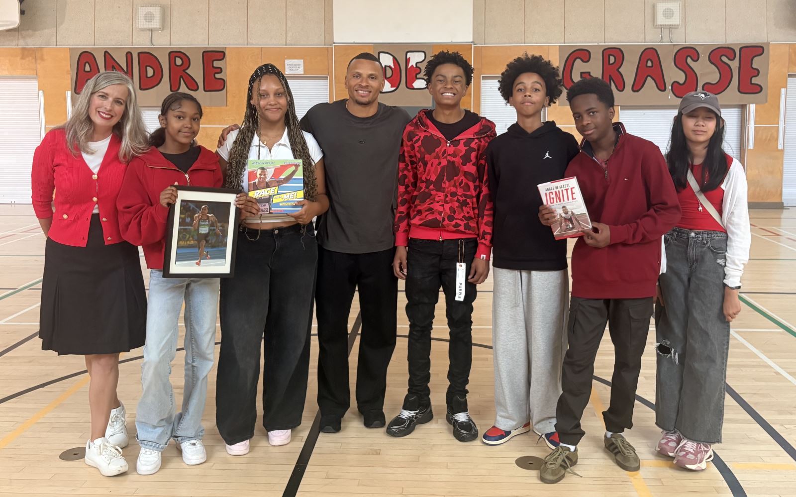A principal stands alongside students and in the centre of the group is Canadian Olympian Andre De Grasse. The group is standing in a gymnasium with a banner in the background with Andre's name painted like the Canadian flag.