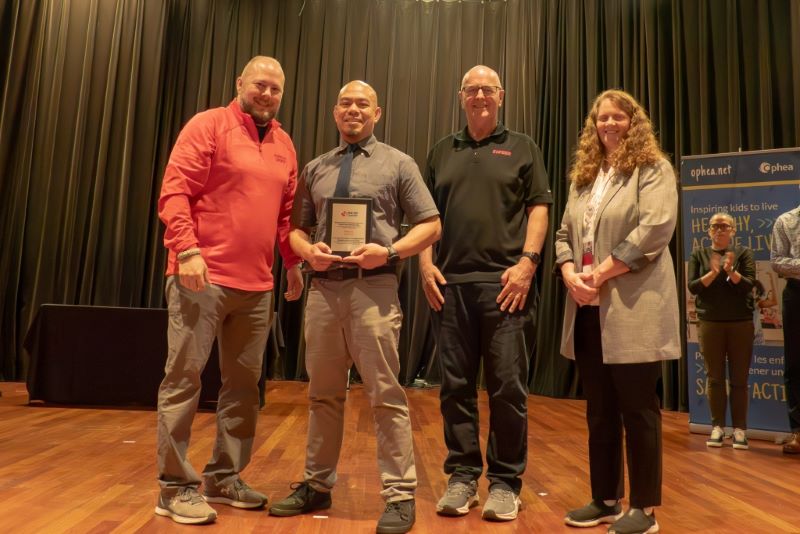Four smiling adults stand in a row side by side on a stage, three men and one woman, the man second from left is holding an award from PHE Canada.
