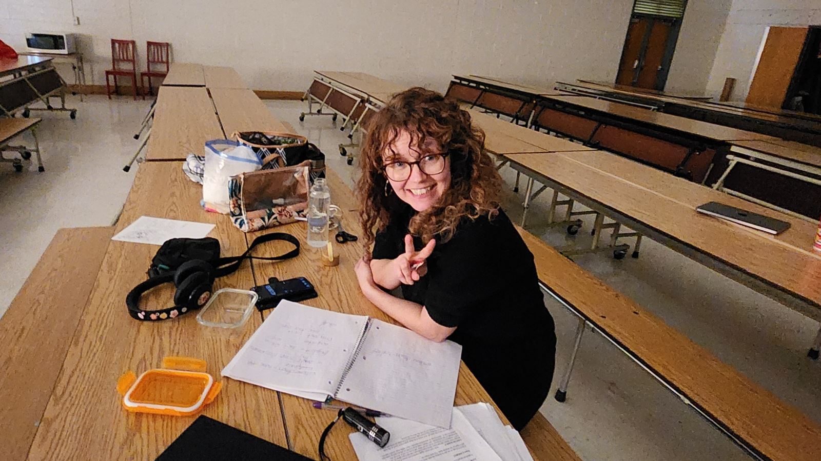 Lisa sits at a cafeteria table reviewing notes and a script during rehearsal, with notebooks, headphones, and personal items spread out in a large multipurpose room.