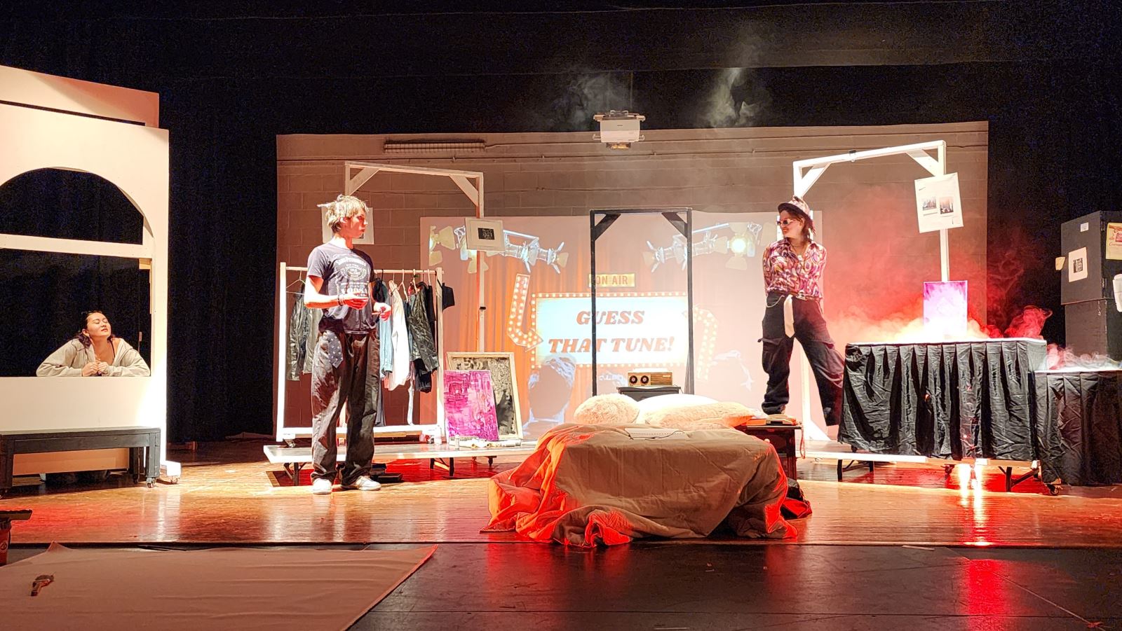 Students perform a scene from a theatre production on stage, surrounded by set pieces, costumes, lighting effects, and projected visuals during a live performance.