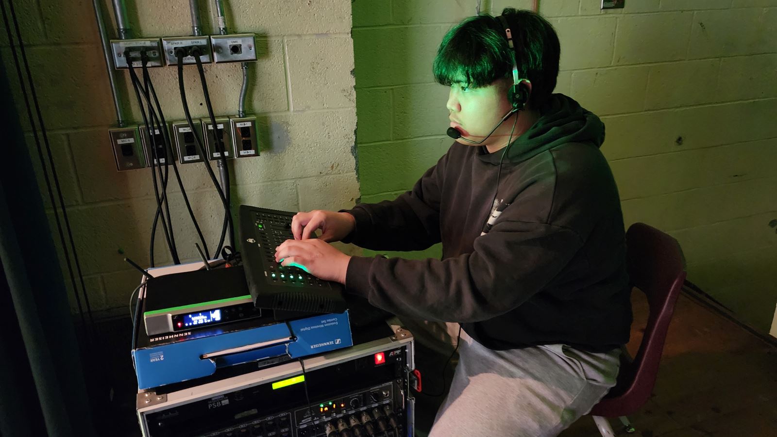 Student operates sound and technical equipment backstage during a theatre production, adjusting controls while wearing a headset.