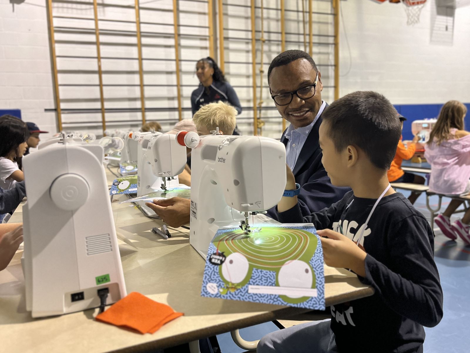 A group of children and an adult are seated at a long table using sewing machines in a gymnasium.