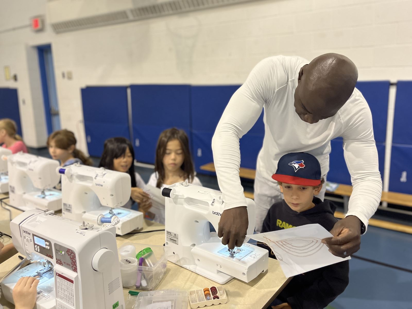 A group of children seated at a table using sewing machines, while a person stands nearby assisting one of them.