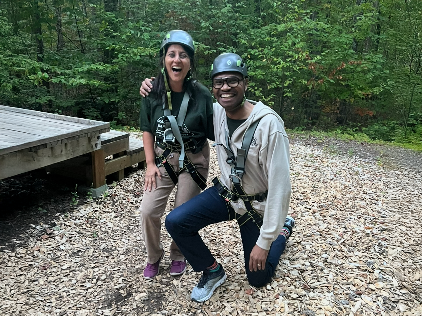 Two people wearing helmets and harnesses pose together in a wooded area.