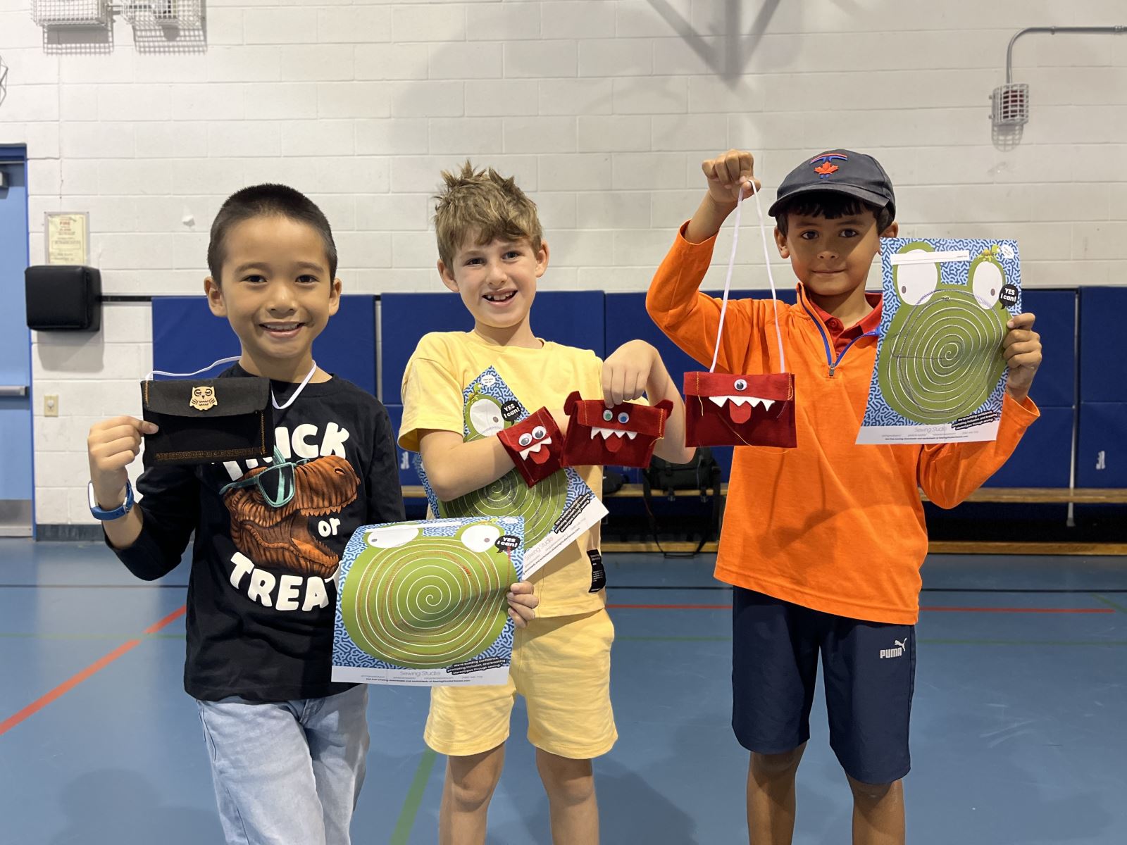 Three children standing in a gymnasium, each holding up a craft project.