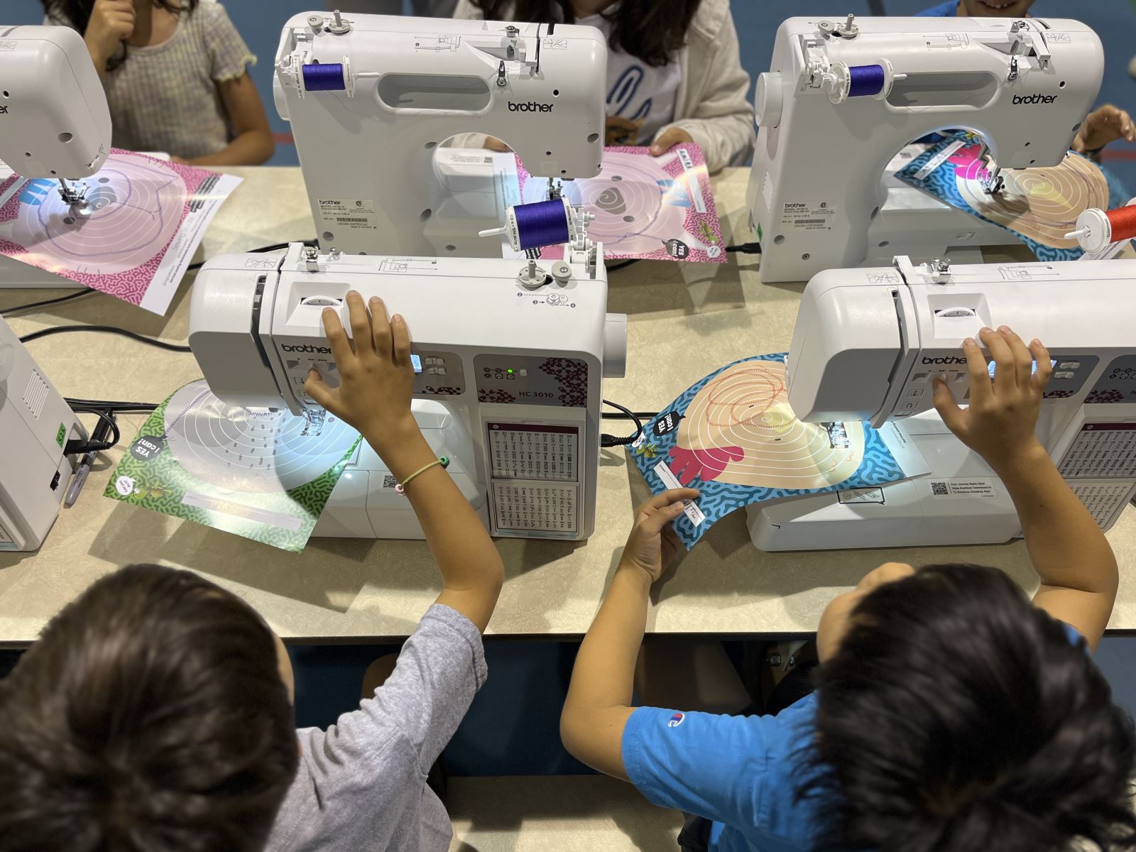 A group of children seated around a table, each using a Brother sewing machine to work on colourful fabric pieces with circular patterns.