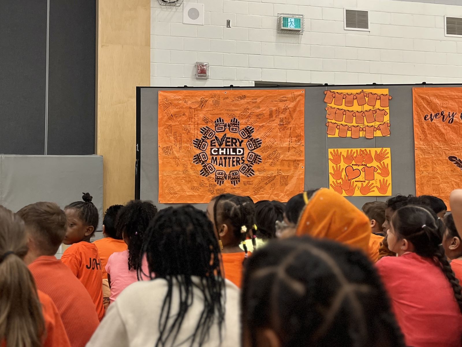 A group of children sit facing a display of orange banners in a gymnasium. The central banner reads "Every Child Matters," surrounded by handprints and other designs.