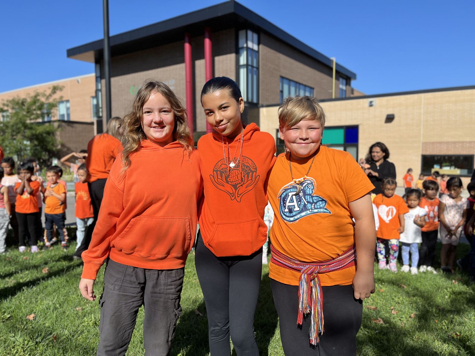 Three children stand together outside on a sunny day, all wearing orange shirts. They are in front of a school building with other children and adults visible in the background, also dressed in orange.