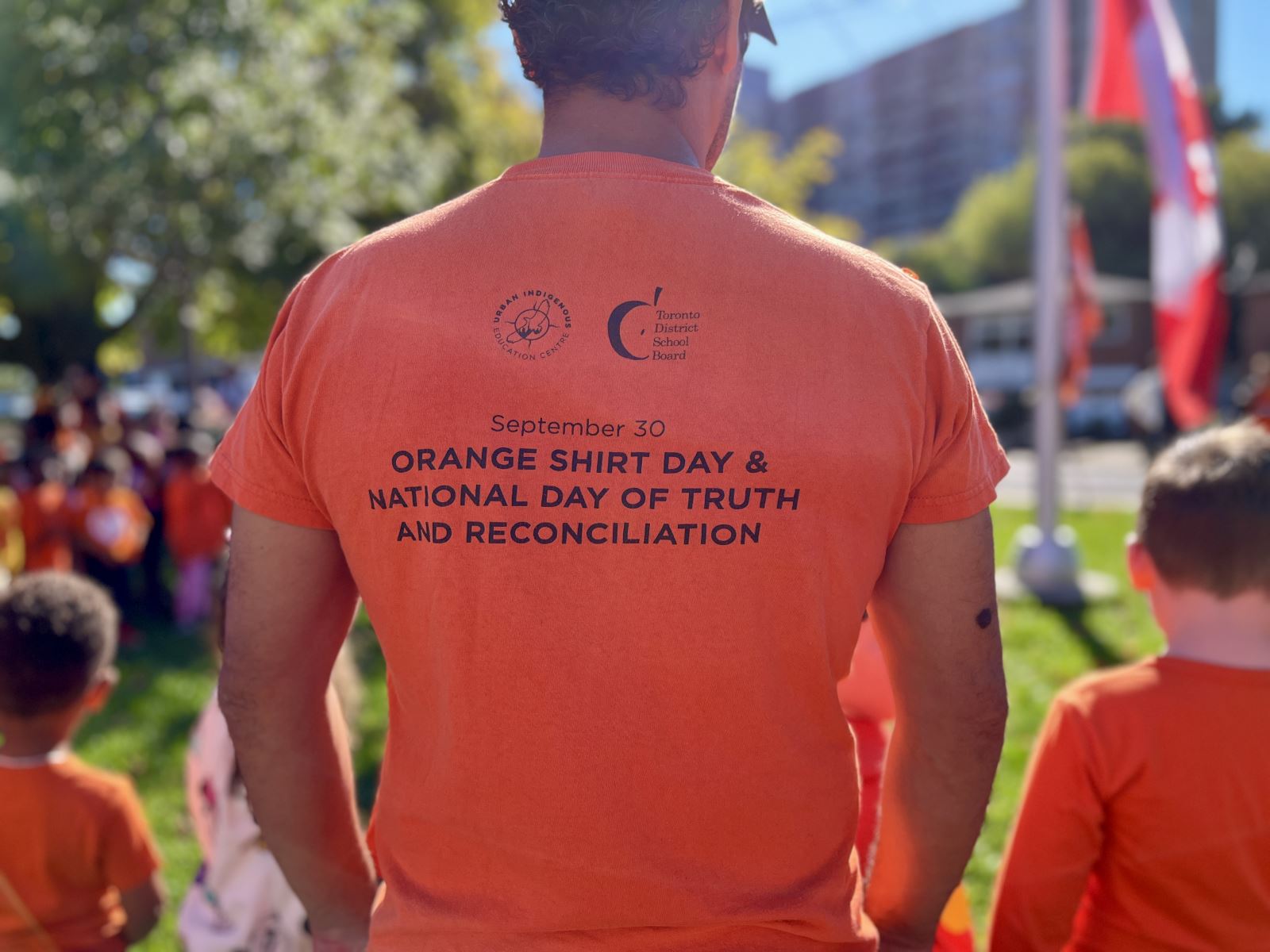 A person wearing an orange shirt with text on the back that reads "September 30 ORANGE SHIRT DAY & NATIONAL DAY OF TRUTH AND RECONCILIATION." The person is standing outdoors among a group of people, many of whom are also wearing orange shirts. A Canadian flag is visible in the background.