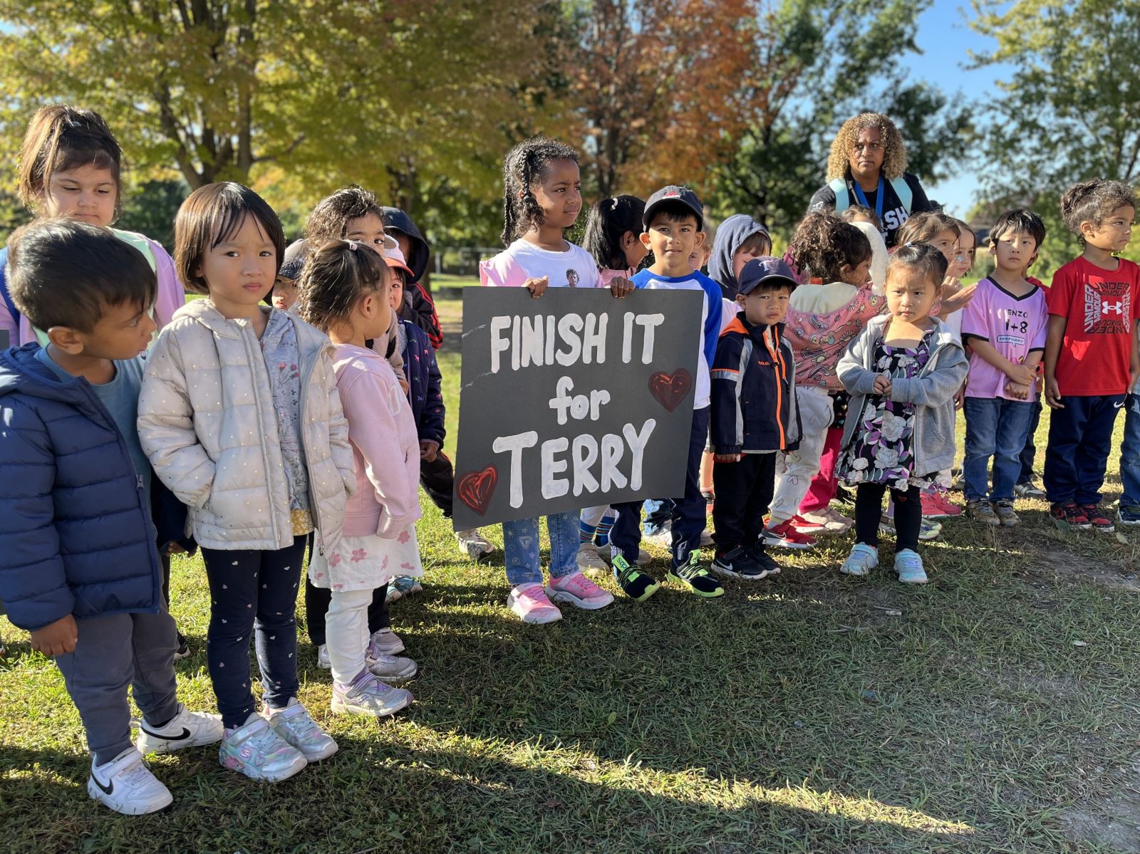 A group of young children standing outdoors on grass in a park, holding a large black sign with white letters that reads “FINISH IT for TERRY” with two red heart drawings. 