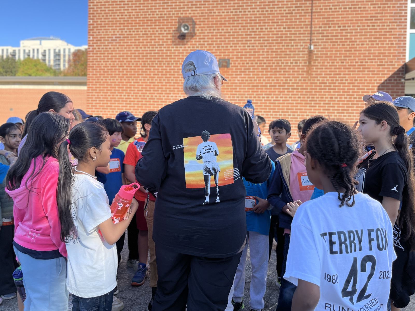 A group of children gathered around an adult wearing a black long-sleeve shirt with an image of Terry Fox running against a sunset background on the back.