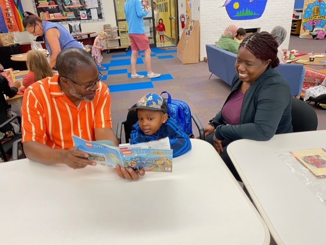A young child sits between two adults at a library table. The child looks at a LEGO book while one adult reads aloud and the other smiles warmly. Other families are reading together in the background.