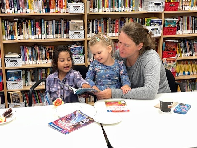 Two young children sit with an adult at a library table, they are looking at a picture book together while surrounded by shelves of books. Plates with snacks and a cup are on the table.