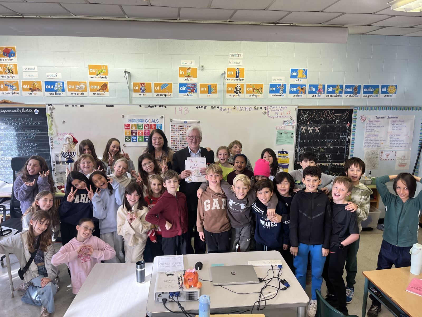 A group of students stands smiling in front of a chalkboard in a classroom with an older woman and older man, former Premier of Ontario Bob Rae, standing at the centre holding a sign that says "thank you for coming"