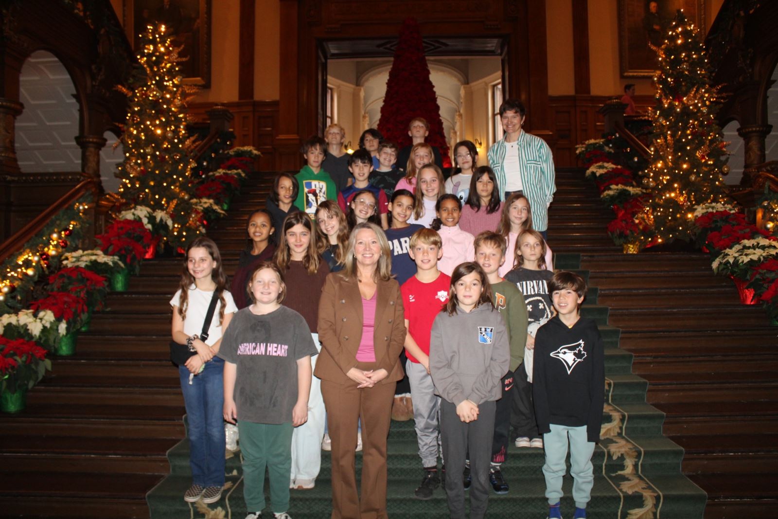 A group of students stands on the steps of Queen's Park, with a woman, MPP Marit Stiles, standing in the centre of the students. The steps are decorated for the holidays on either side with poinsetta plants, lights, and mini Christmas trees.
