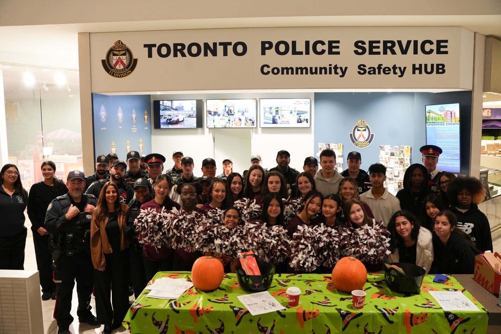 A group of cheerleaders stands surrounded by a group of police officers under the Toronto Police Community Safety Hub sign.
