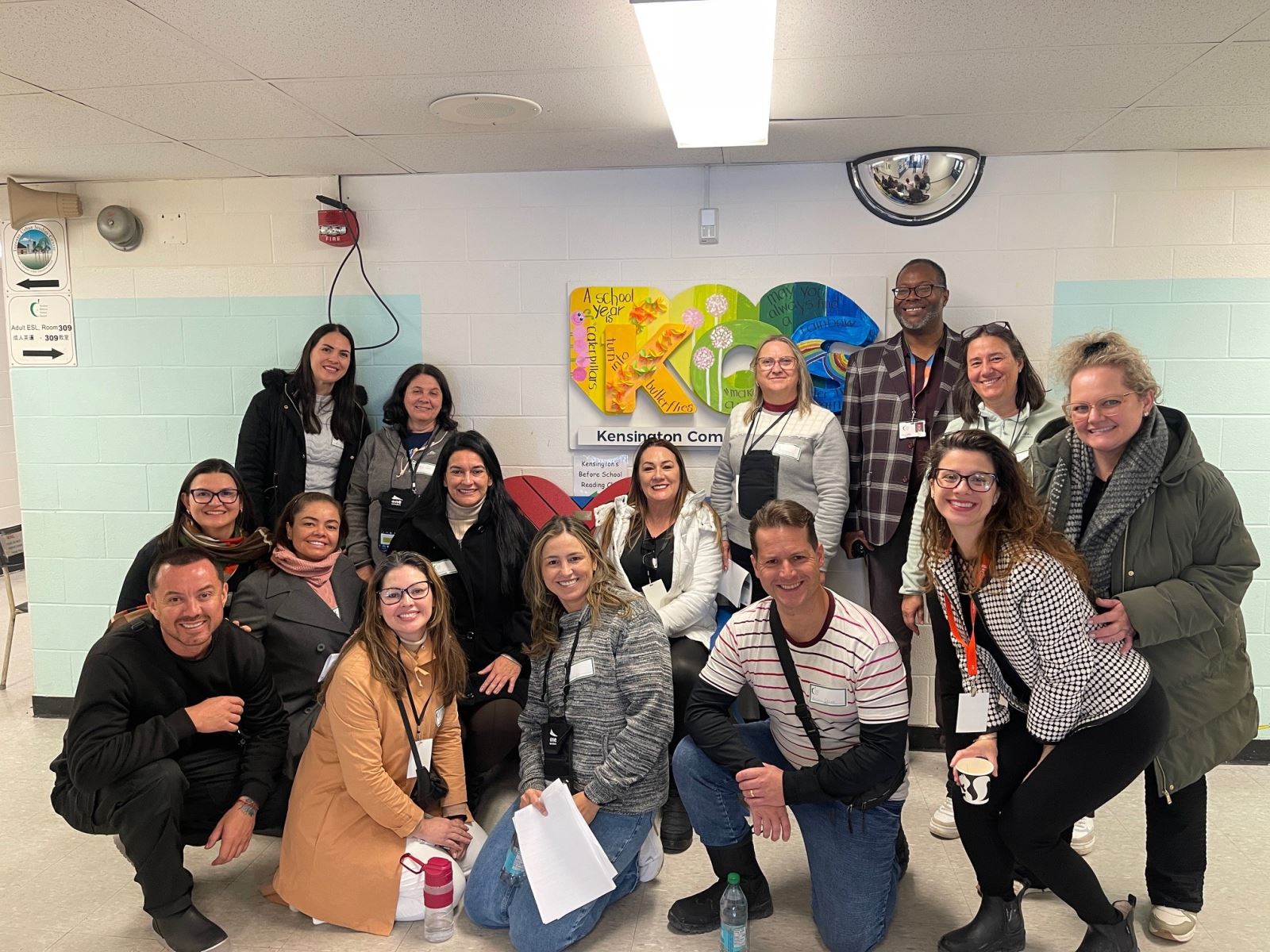 Group of adults smiling and looking at the camera grouped together in a hallway of a school in front of a "KCS" sign for the school name's acronym. 