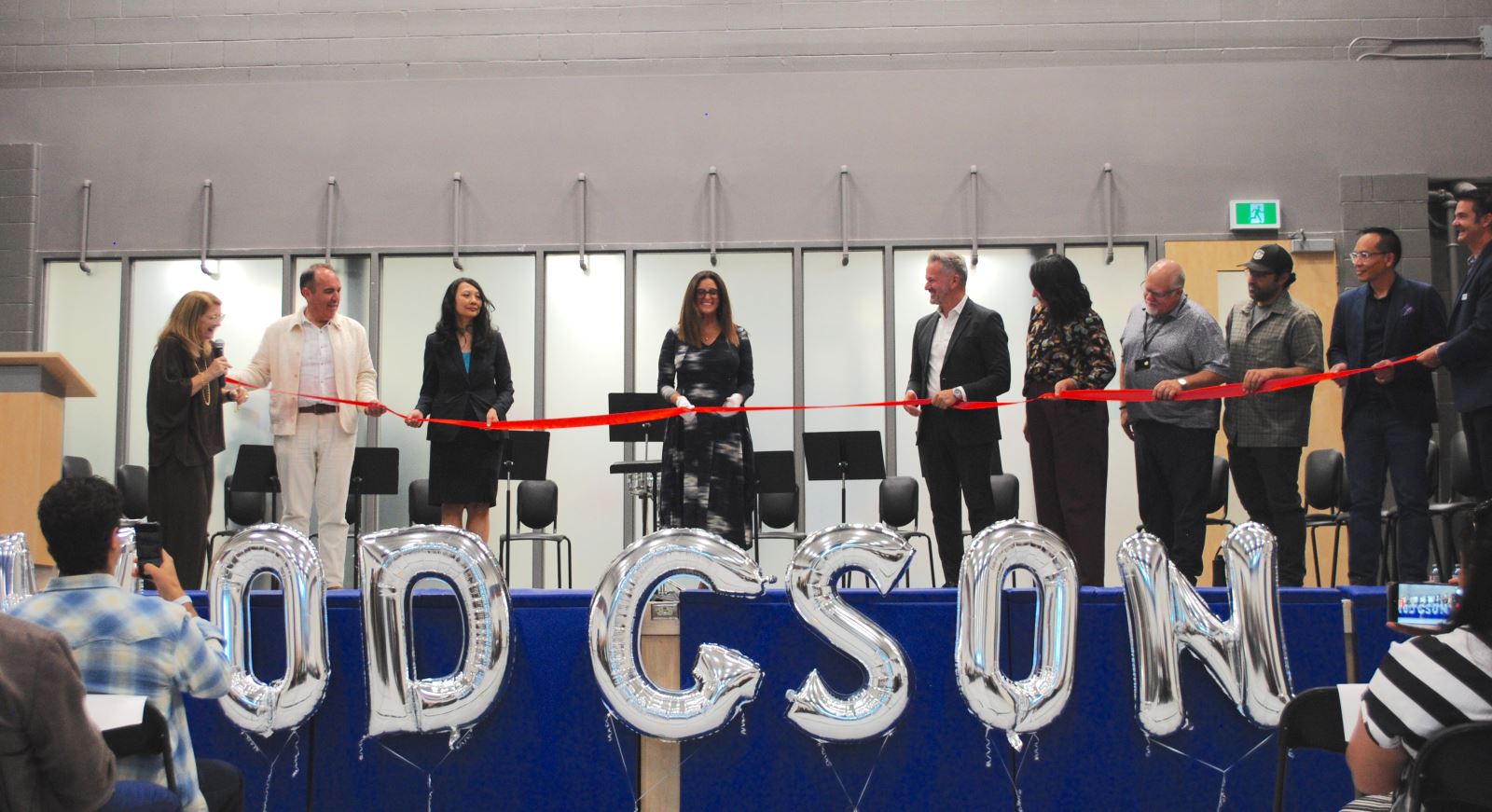 A group stands on a stage holding a red ribbon ready to be cut. At the front of the stage the school name "Hodgson" is spelled out in large bubble letters. 