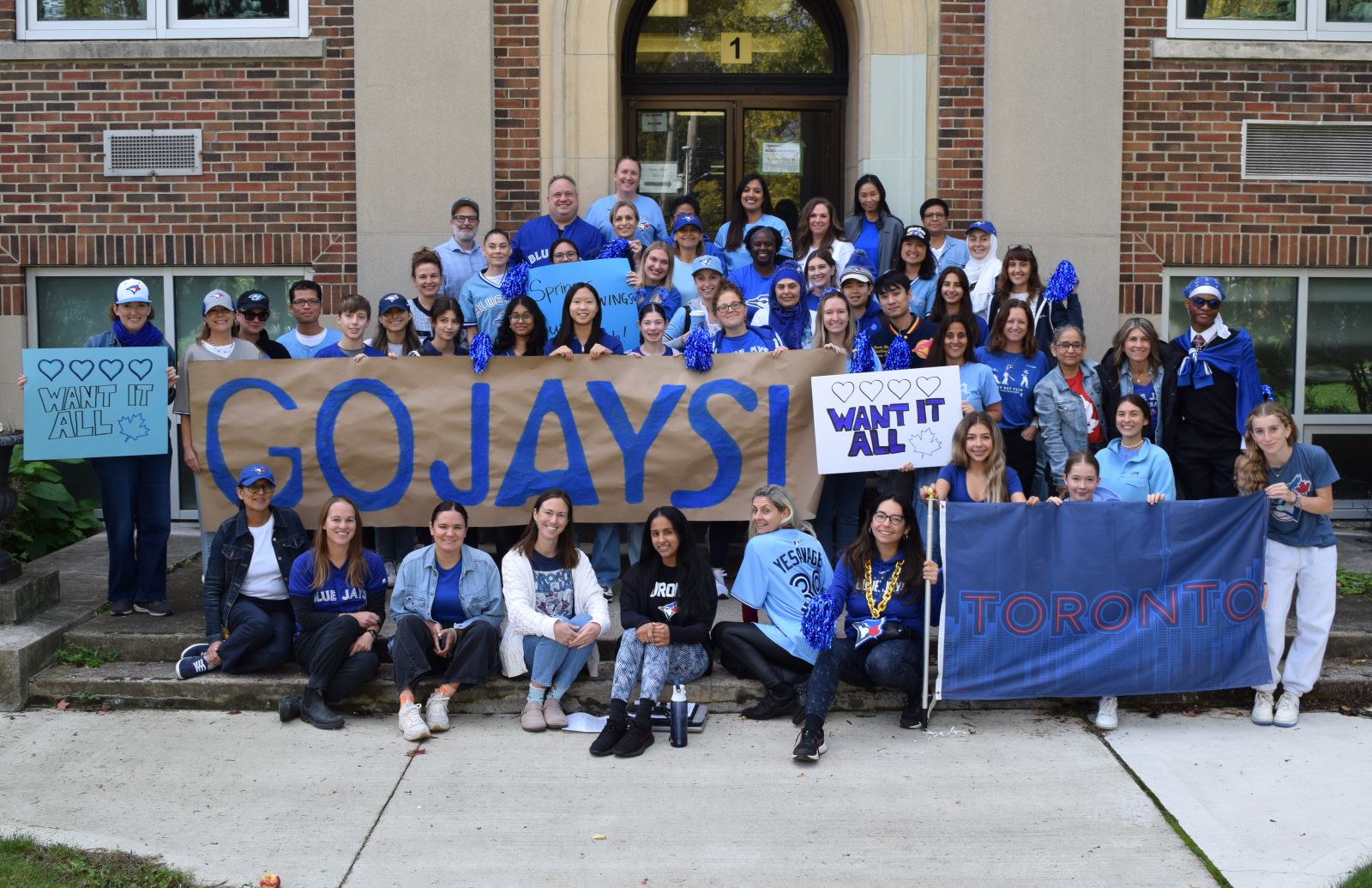 A large group of staff in blue and Blue Jays gear stand in front of a school building holding various painted signs with "Go Jays" and "Want it All!"