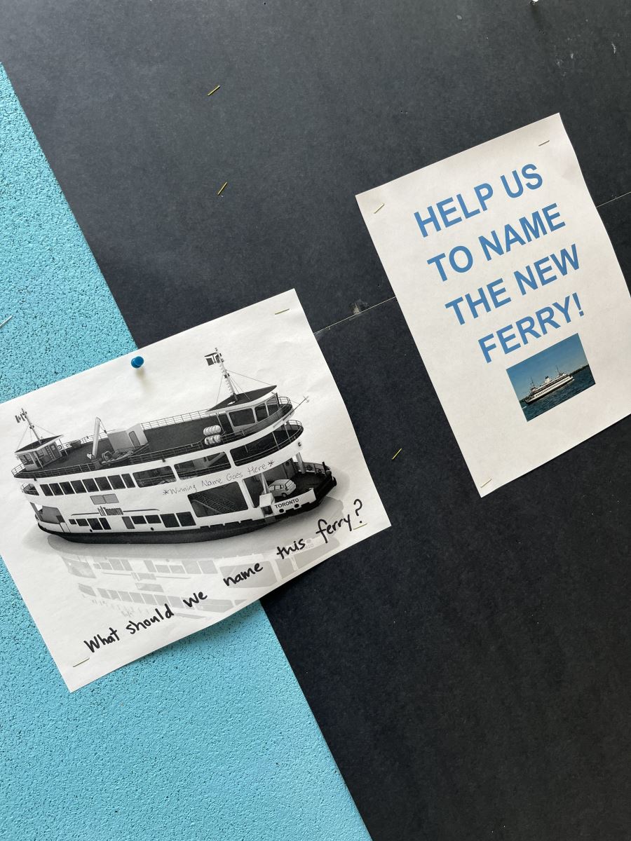 Two posters pinned to the wall, one with the image of a Toronto ferry and "winning name here" written on the front of the boat, and a second poster with text "HELP US NAME THE NEW FERRY"