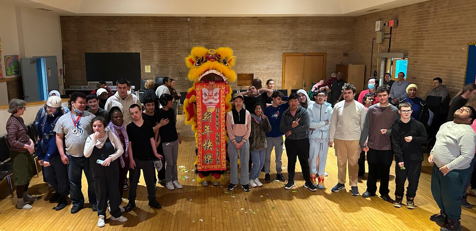 A large costume lion stands in the centre of a room with groups of students standing on either side of the lion for a posed photo.