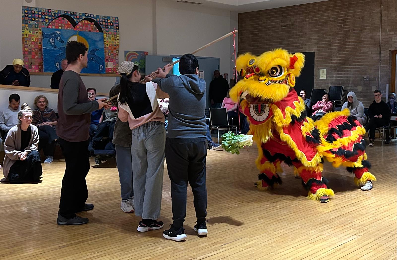 Three students whole a long pole with lettuce tied to the end of it. They are dangling the pole in front of a large costume lion who is looking at the lettuce ready to eat it.