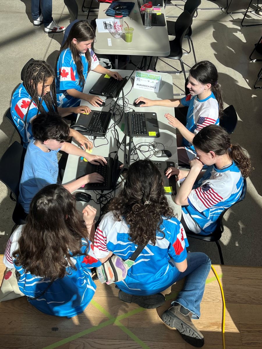 Students wearing matching shirts sit at a group table each focused on the screen of a laptop in front of them 