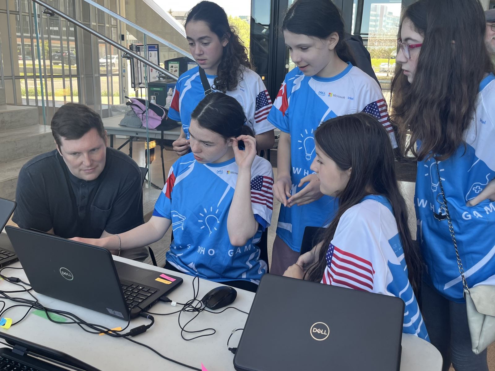 A group of students in the same shirt and one adult crowd around the screen of a computer.