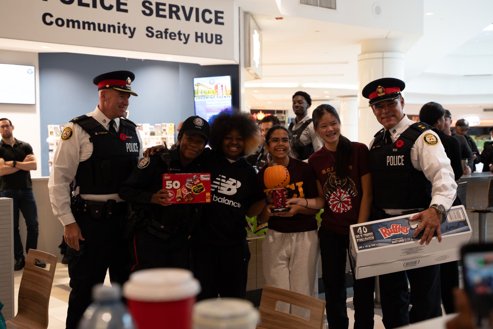 Group of students holding pumpkins and candy stand in a row with police officers on either side in a congratulatory pose, the group stands under a Toronto Police Community Safety Hub sign.