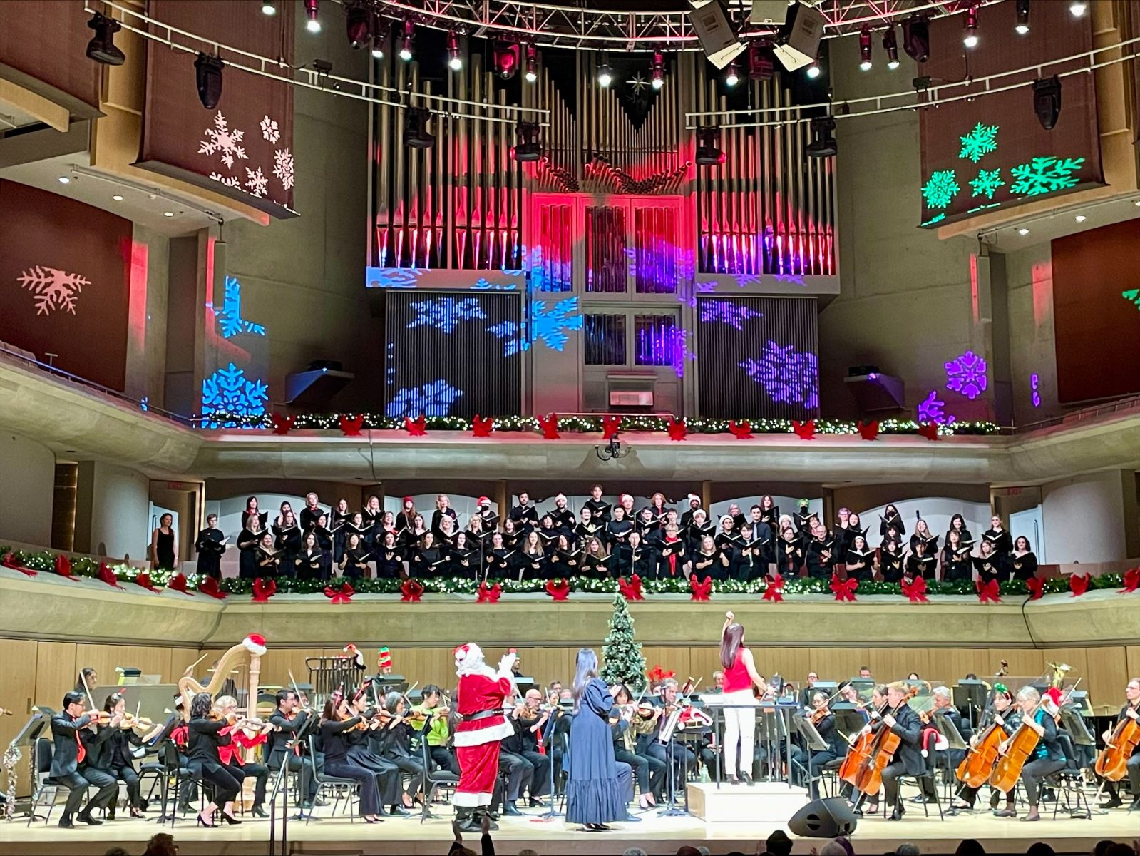 A choir stands on the balcony looking down over a stage where the Toronto Symphony Orchestra sits with Santa Claus standing at the front of the stage applauding the choir.