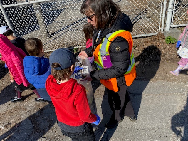 A group of students surround a staff member in a vest holding a clear garbage bag, students are placing garbage in the bag.