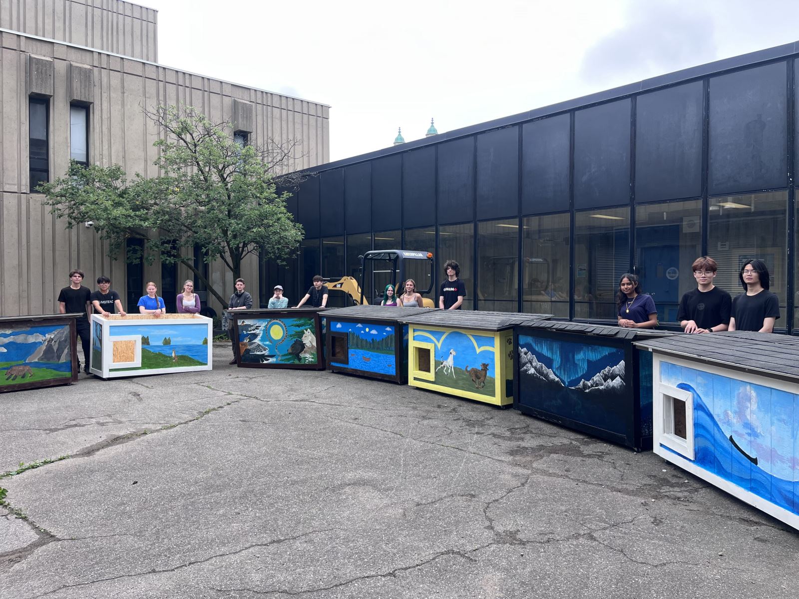 A row of dog houses that are decorated with colorful murals of nature scenes and animals sits lined up in front of students who stand behind the boxes proudly displaying their finished work.