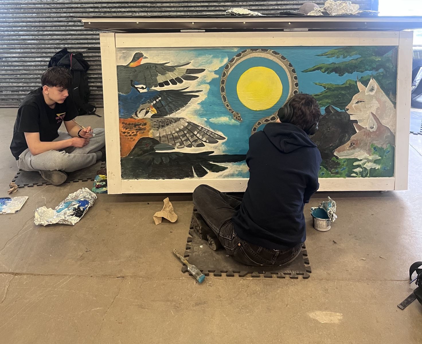 Two students sit on the ground beside a completed dog house painting a colorful and detailed mural. 