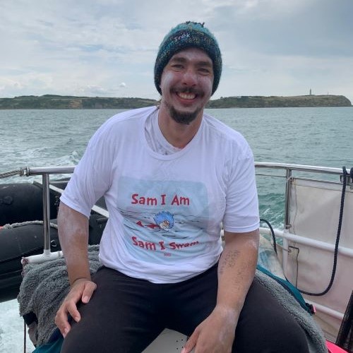 A smiling man sits on a boat after a swim, wearing a knit beanie and a white T-shirt that reads “Sam I Am, Sam I Swam” with a cartoon swimmer on it. His arms are covered in white zinc sunscreen, and handwritten notes are visible on one forearm. The sea and a distant coastline with cliffs and a lighthouse appear in the background under a cloudy sky.