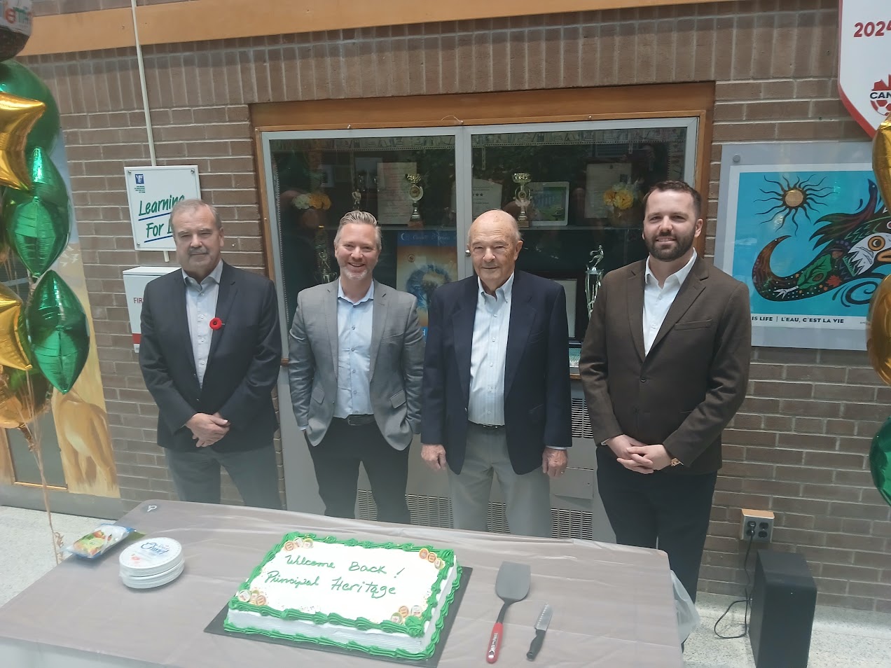 Four individuals standing behind a table with a large rectangular cake decorated in green and white icing that reads “Welcome Back Principal Heritage.” Green and gold balloons are visible on both sides.