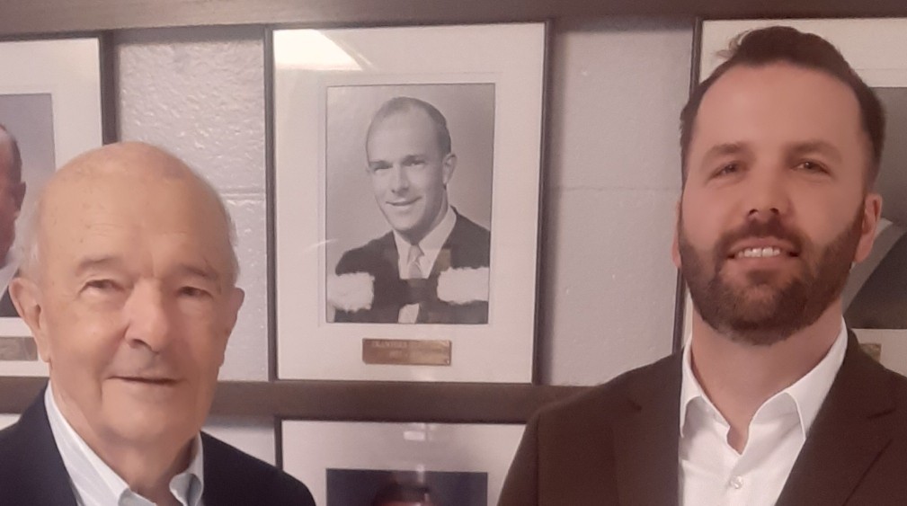 Two individuals standing in front of a wall displaying framed portraits of former principals, including a photo labeled “Crawford Heritage.”
