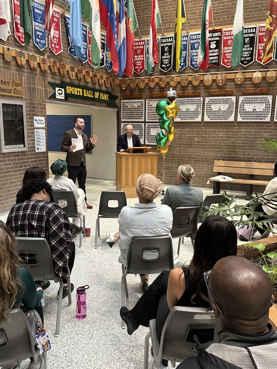 Two individuals at the front of a school hallway, one speaking and one standing at a podium, addressing a seated audience. Green and gold balloons are visible near the podium, and sports banners hang overhead.