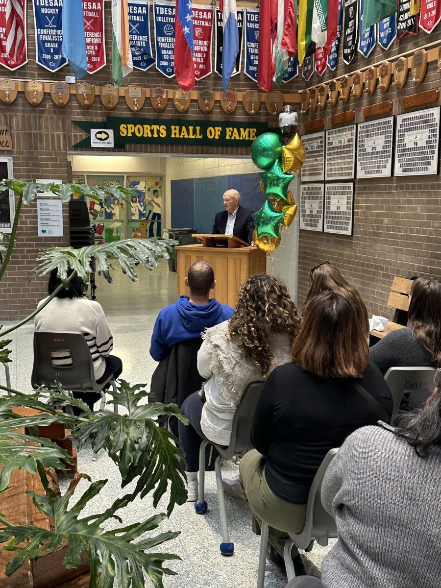An individual standing at a wooden podium addressing a seated audience in a school hallway decorated with sports banners and green and gold balloons.
