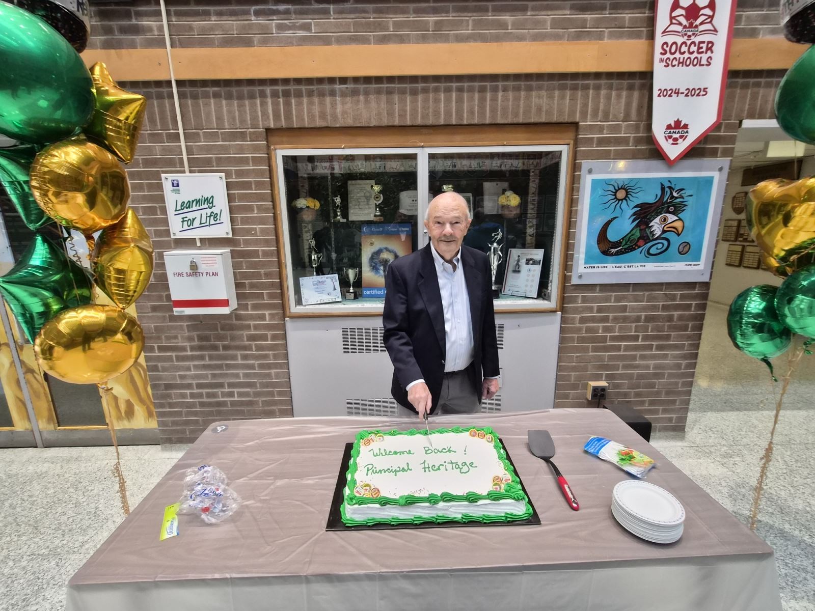 An individual standing behind a table with a decorated cake that says “Welcome Back Principal Heritage,” holding a knife as if preparing to cut the cake. Green and gold balloons are arranged to the side.