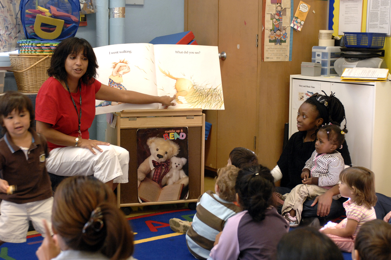 Educator pointing to book with students and parents on carpet listening