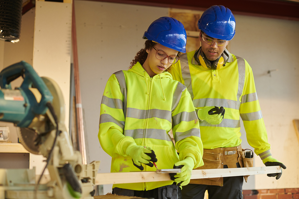 Student measuring plank of wood, dressed in reflective jacket, and educator in reflective jacket observes