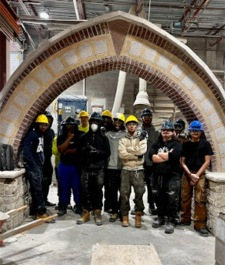 A dozen students in hard hats pose under stone archway