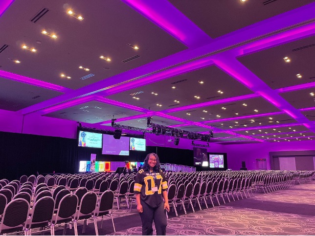 Student stands and poses for photo in empty conference room with rows of empty seats